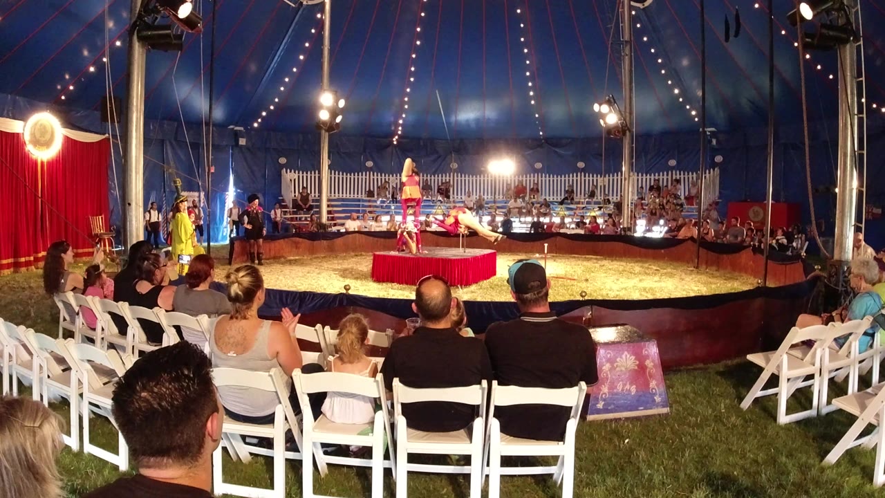 In Broomfield, Colorado, pink-dressed performers stand on and near a red platform inside a blue tent while spectators sit in white folding chairs in the foreground.