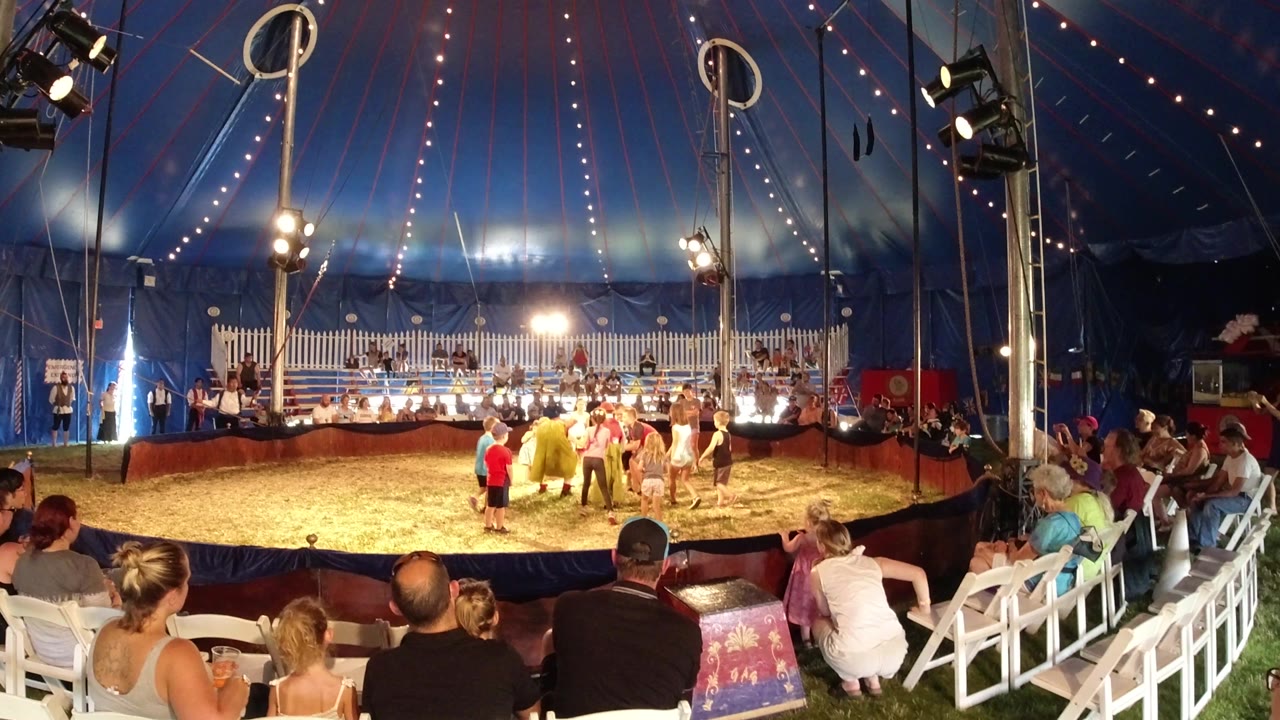 A performer in a yellow costume stands near children in the Broomfield circus ring as the audience watches from white chairs around the tent edge.