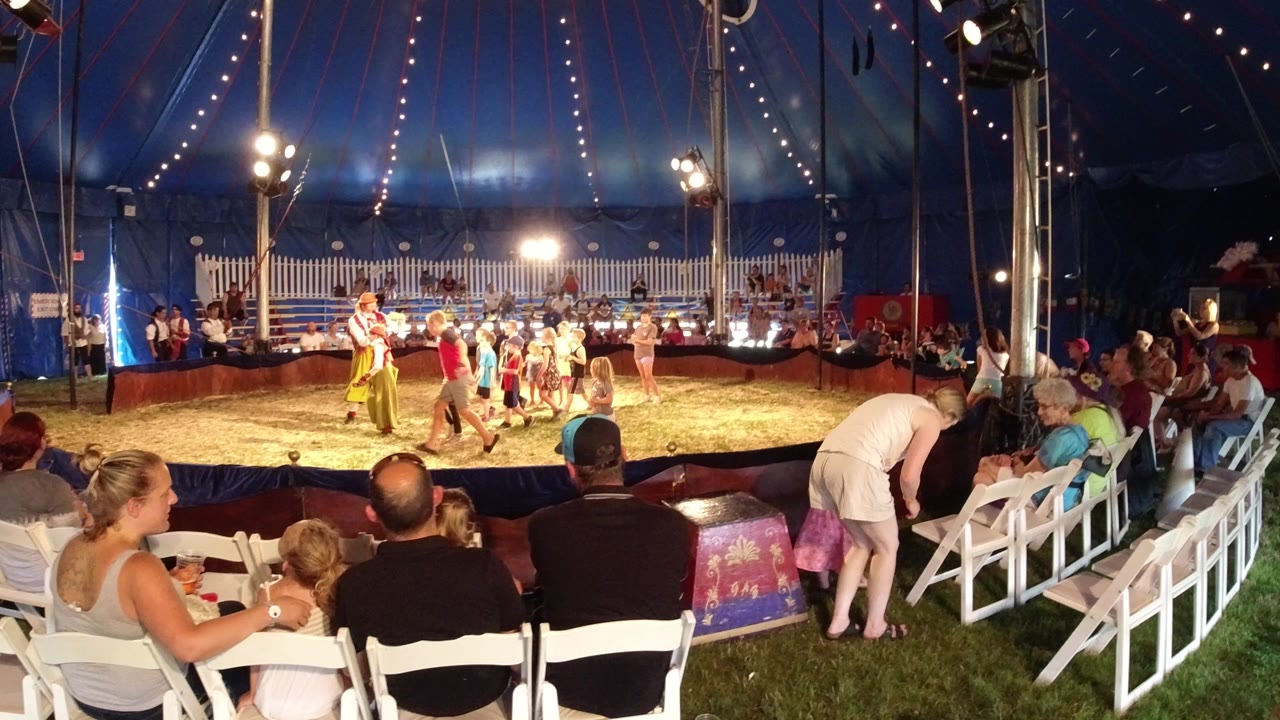 Wide view of children in a tent ring with spectators seated in white chairs.