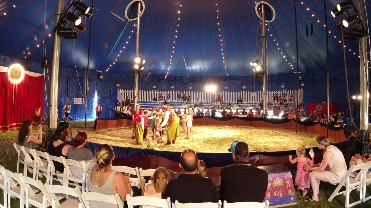 Families seated in a circus tent watch children and adults near a clown in the ring.