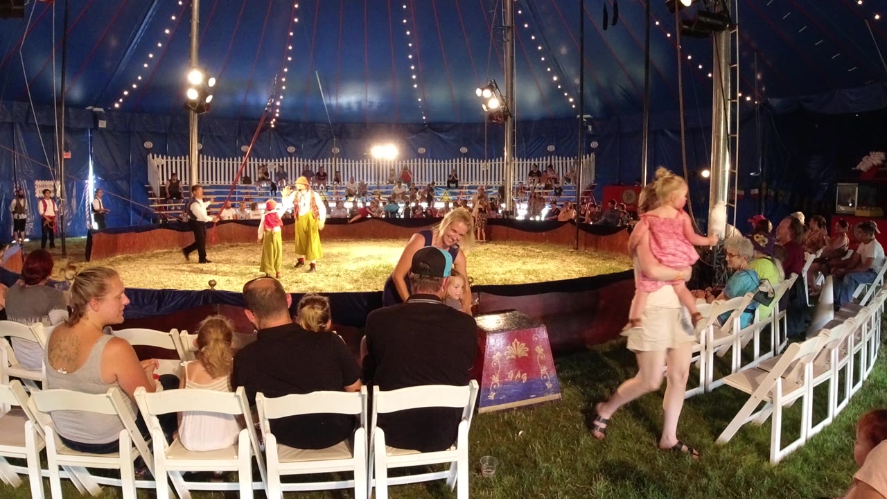 Broomfield, Colorado circus scene with spectators seated in white chairs, a girl near the aisle, and performers under strong lights at the center of the tent.