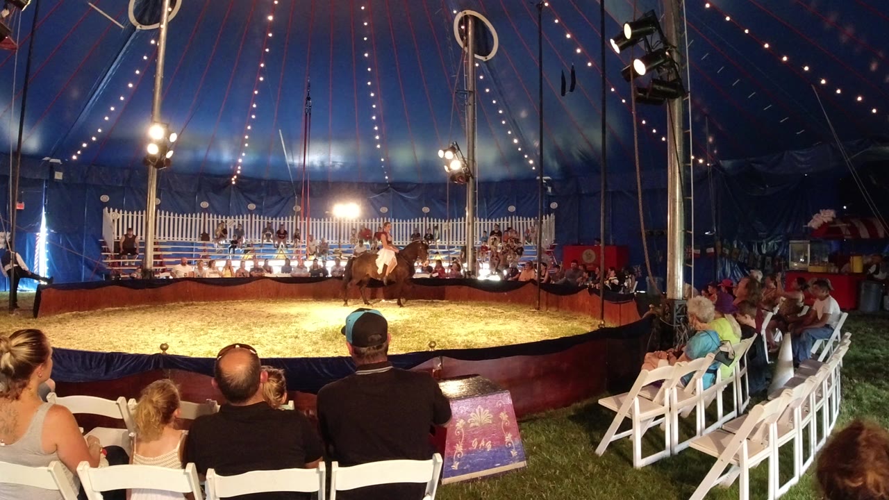 A wide view in Broomfield, Colorado shows a young circus rider on a dark horse near the center of a lit tent ring, with seated spectators in the foreground.