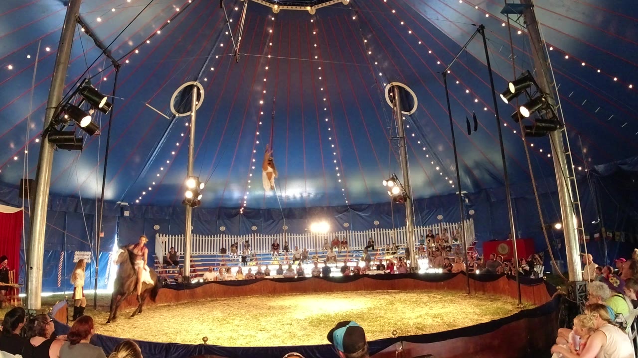 Wide view inside a Broomfield, Colorado circus tent shows an aerial performer suspended high above the ring while a horse and rider move below and spectators watch from the edges.