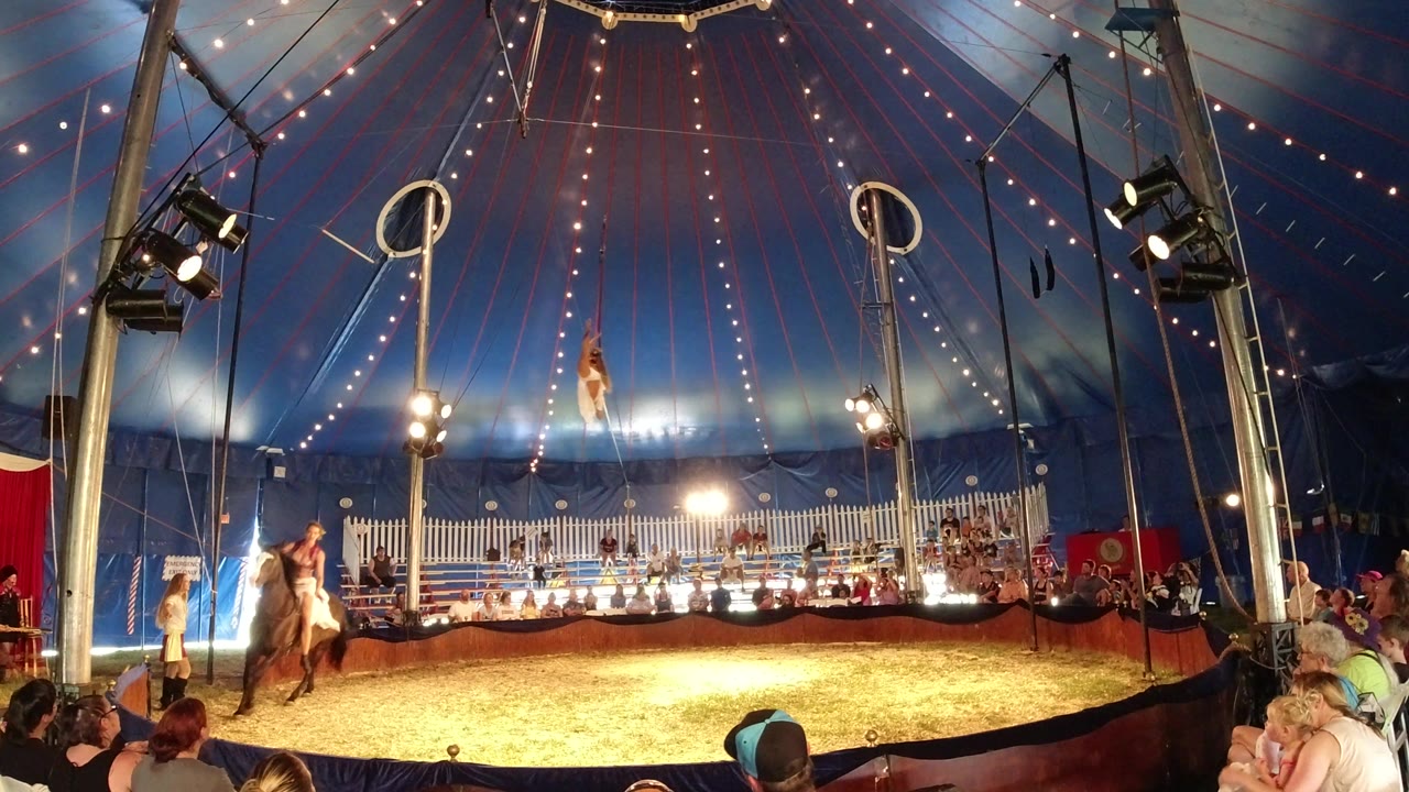 The Broomfield, Colorado circus ring remains in a wide view as the aerial performer hangs near center and the horse rider passes along the left side of the lit sawdust floor.