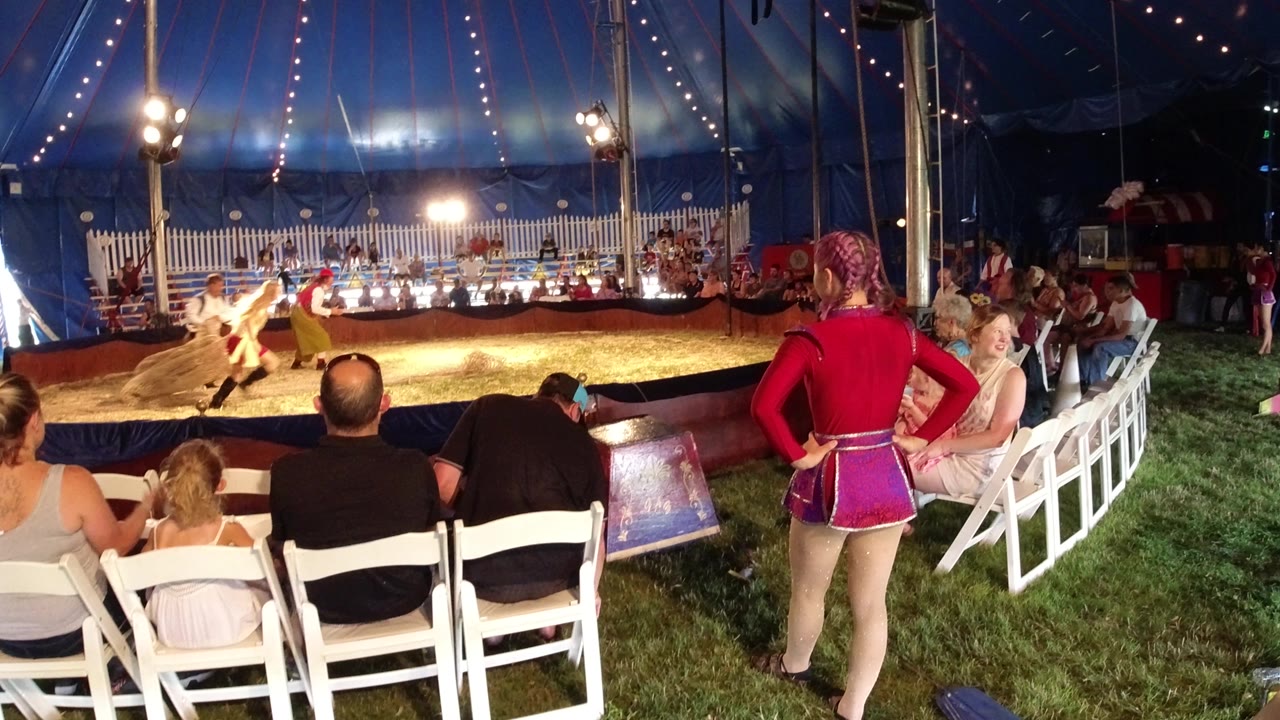 In Broomfield, Colorado, the camera looks past seated spectators toward performers crossing the sawdust ring, with a red-uniformed attendant standing near the right aisle.