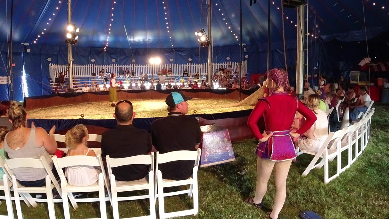 In Broomfield, Colorado, an audience sits on white folding chairs under a blue circus tent while a performer stands in the lit ring near a white fence backdrop.