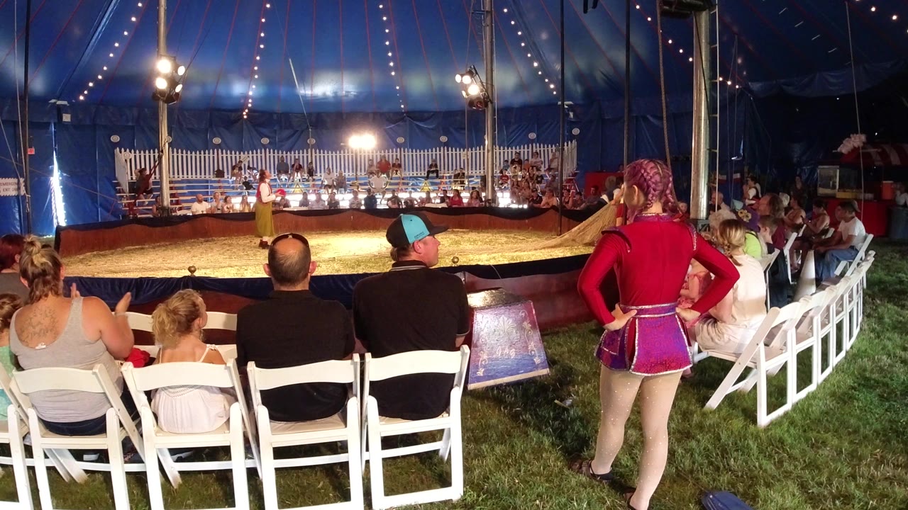 In Broomfield, Colorado, the seated crowd faces the circus ring as a costumed woman stands near the right aisle and the performer remains under bright tent lights.