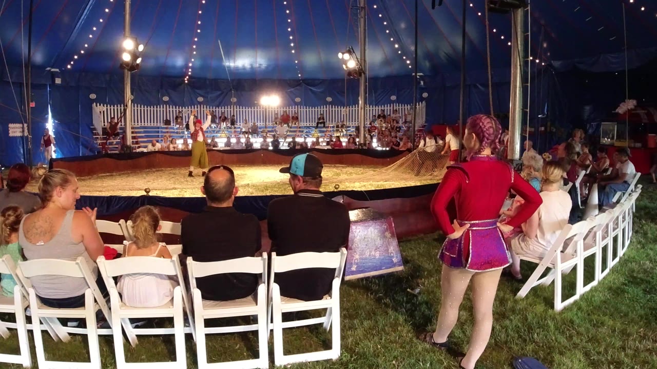 In Broomfield, Colorado, adults and children sit in white folding chairs inside a blue circus tent while performers stand across the ring under bright lights.