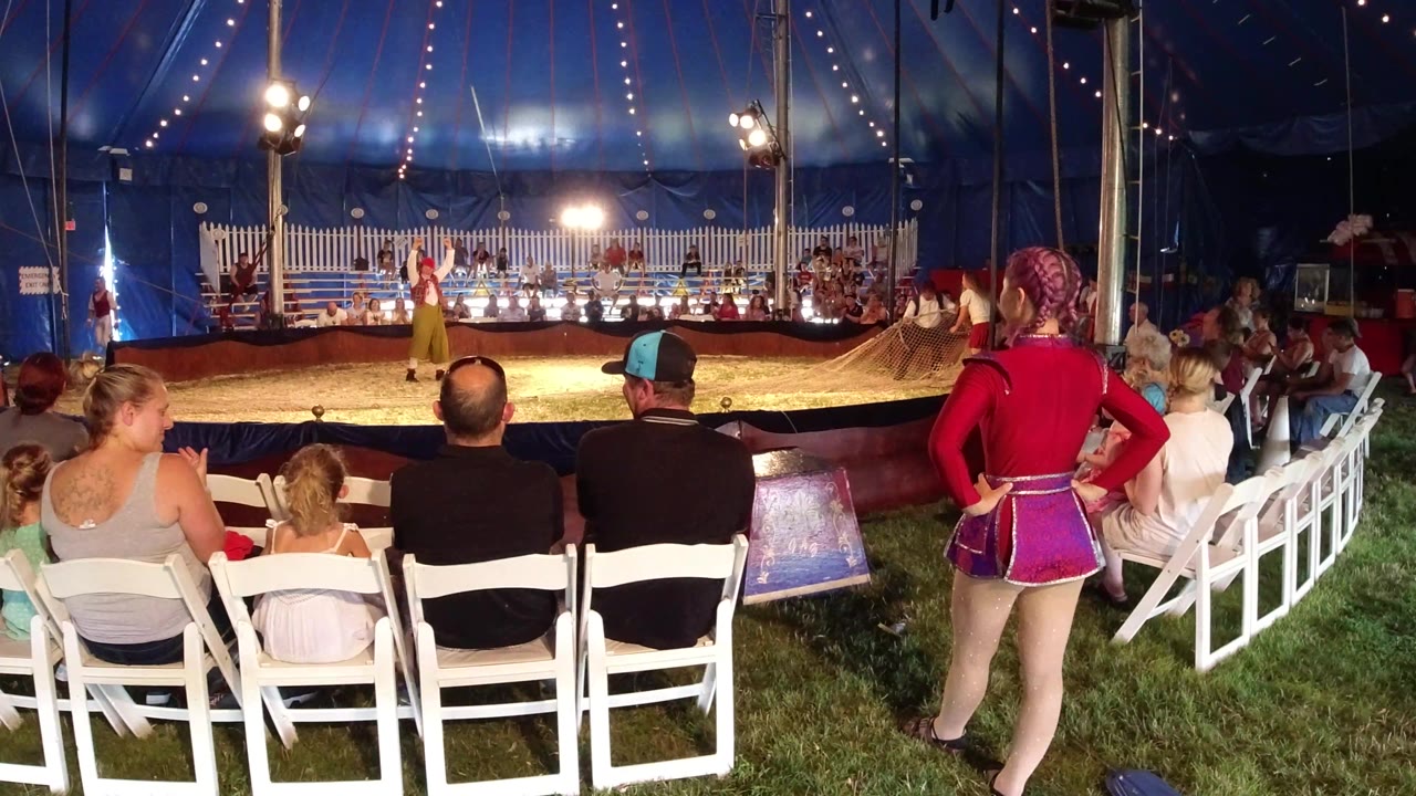 A red-costumed person stands at the right edge while families seated in white chairs watch the lit circus ring in Broomfield, Colorado.
