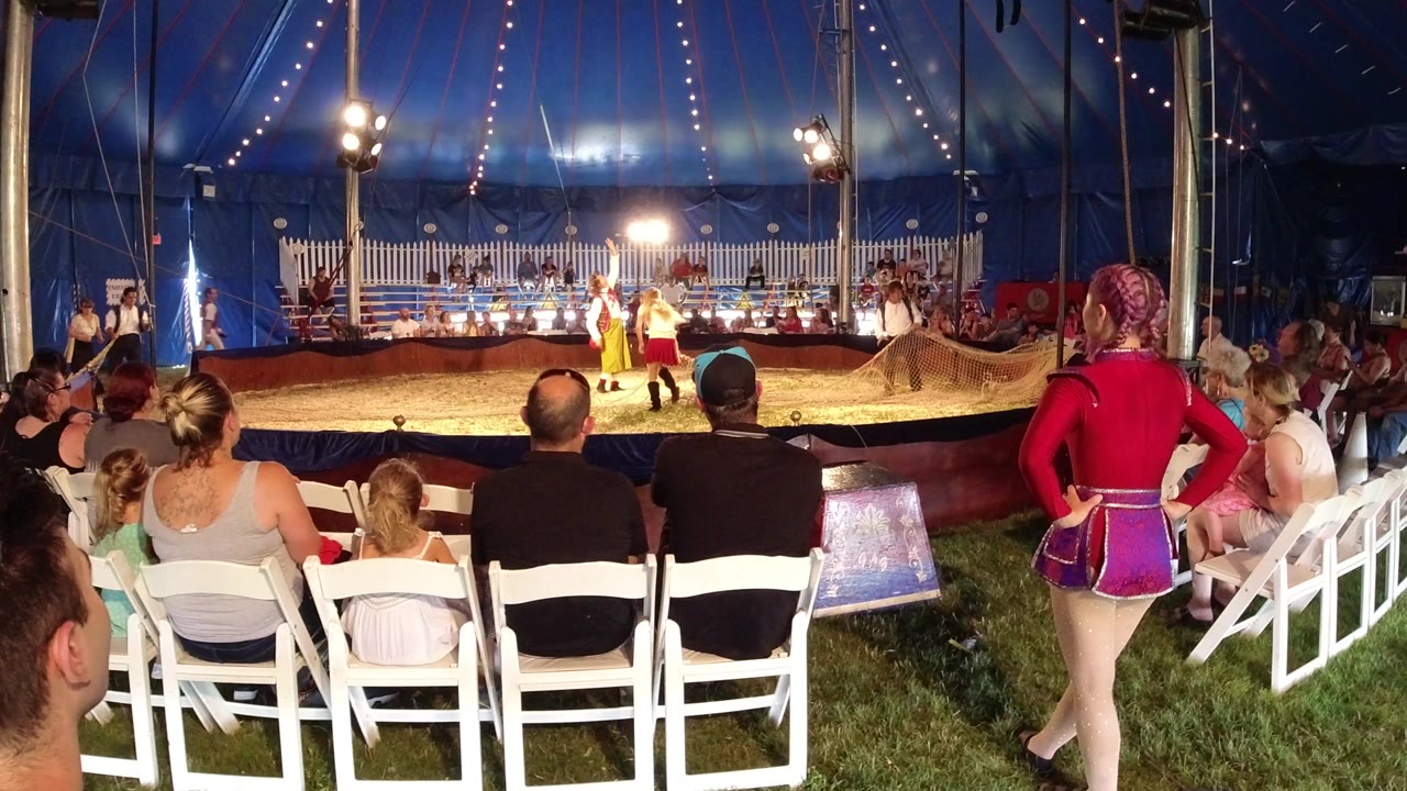 Audience seated on white chairs watching circus performers inside a blue tent.