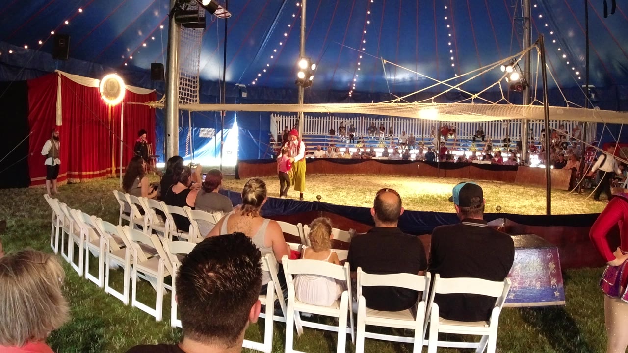 Audience seated in white chairs facing a lit tent performance area.