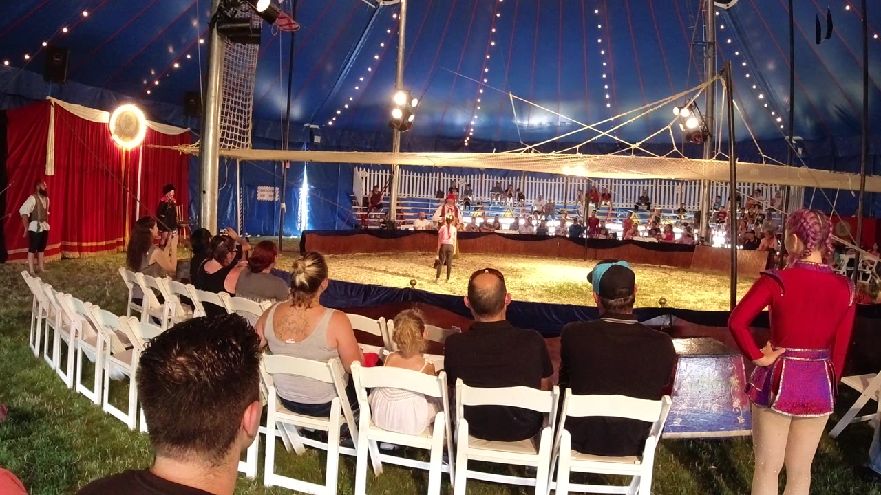 At 0:01, the clown act continues in the ring with the audience in the foreground, a performer in red standing at right, and tent lights glowing overhead in Broomfield, Colorado.