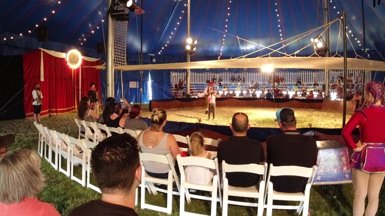 Audience seated in white chairs watching clowns inside a circus tent.