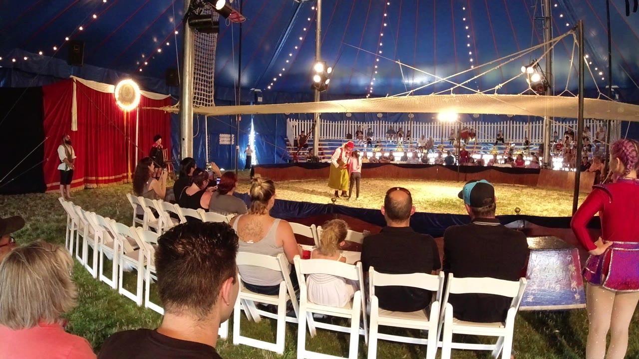 Audience seated in white chairs inside a circus tent watching a performer.