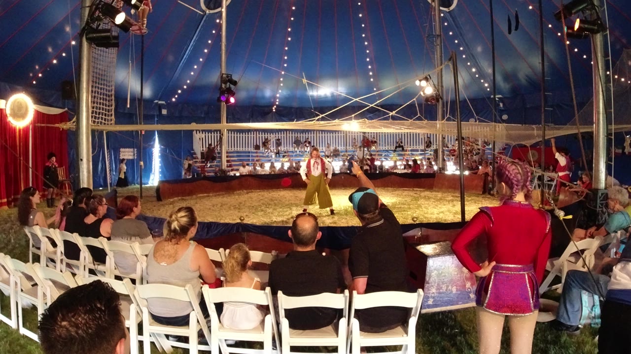 Broomfield, Colorado tent scene with the performer centered in the ring, audience members in the foreground, red side curtains, and bright lamps aimed across the performance area.