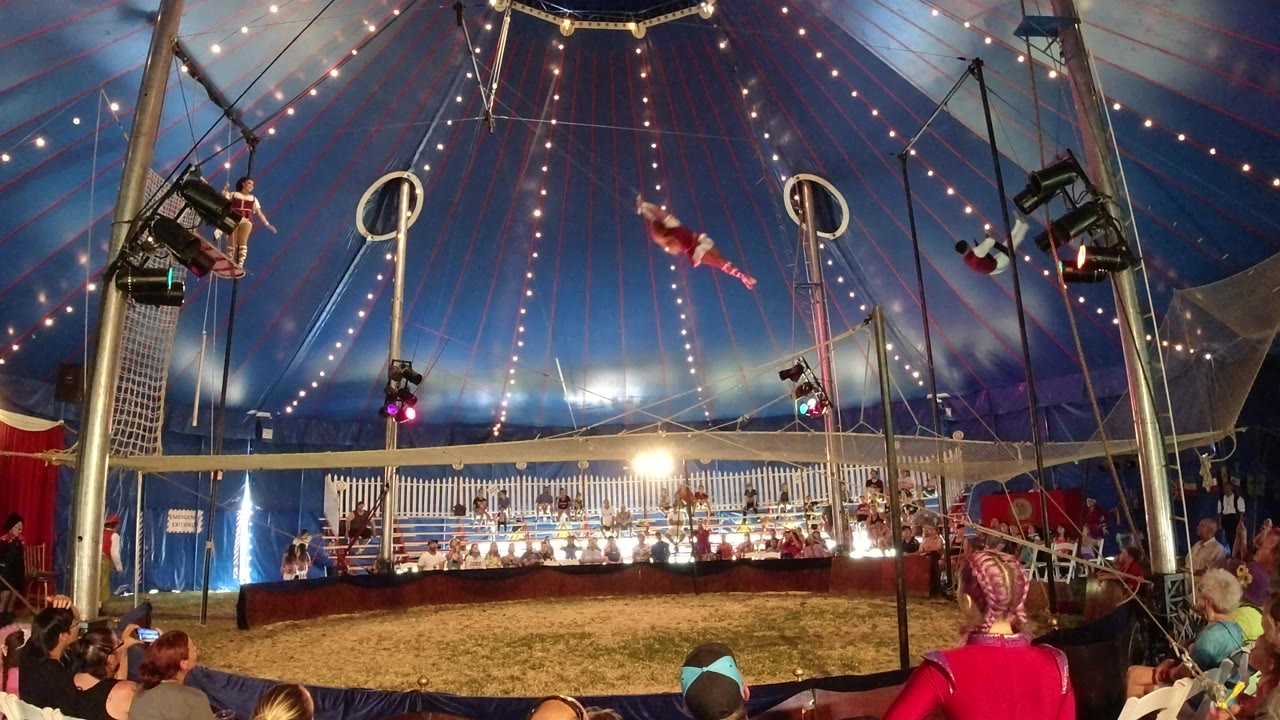 A performer in red swings through the center of the Broomfield circus tent while another performer and audience members remain visible near the rigging.