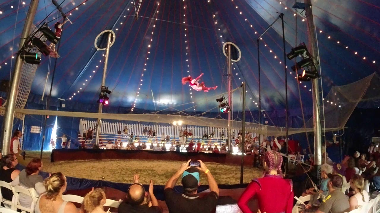 A red-costumed performer hangs near the center of the Broomfield circus tent while spectators in foreground seats look toward the ring.