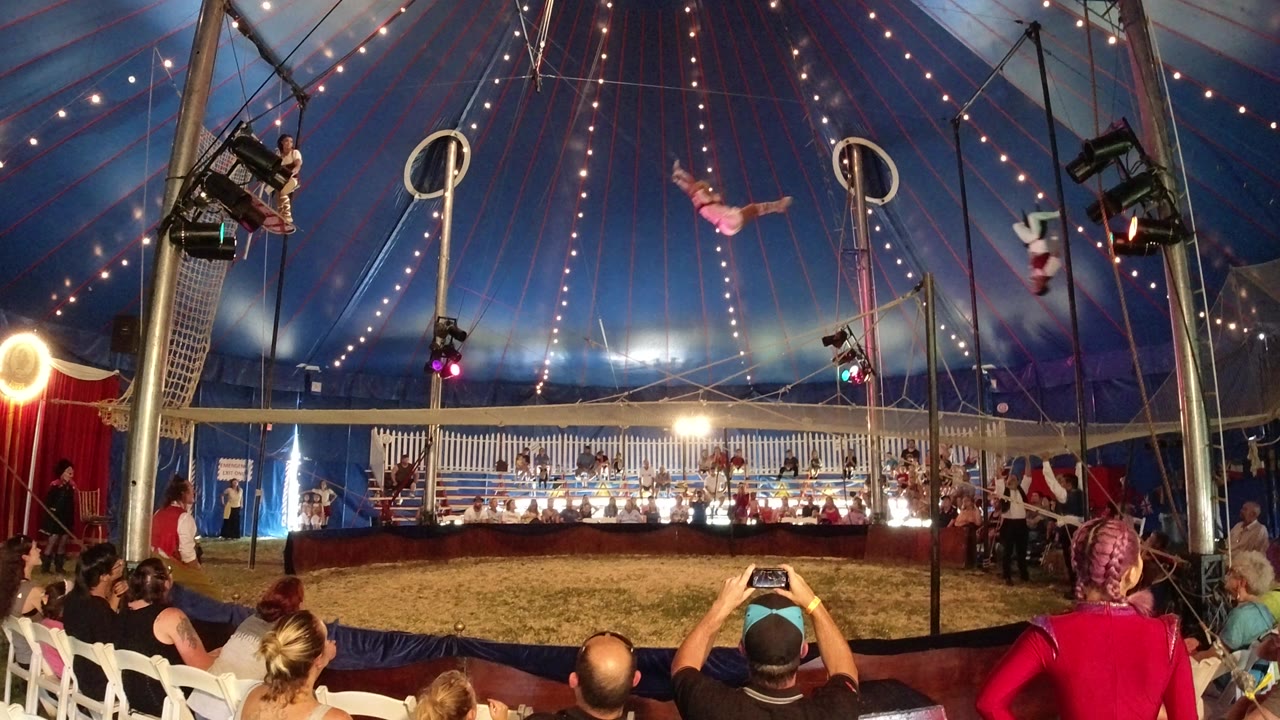 In Broomfield, Colorado, a trapeze performer in pink crosses the center of the tent as other performers remain near the rigging and audience members look on.