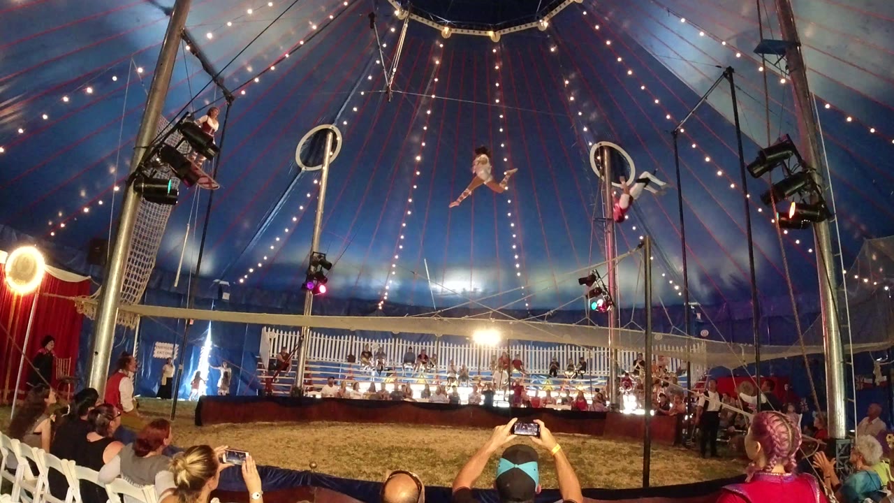 Wide view inside a Broomfield, Colorado circus tent as acrobats perform above a seated audience under strings of warm lights at the start of the clip.