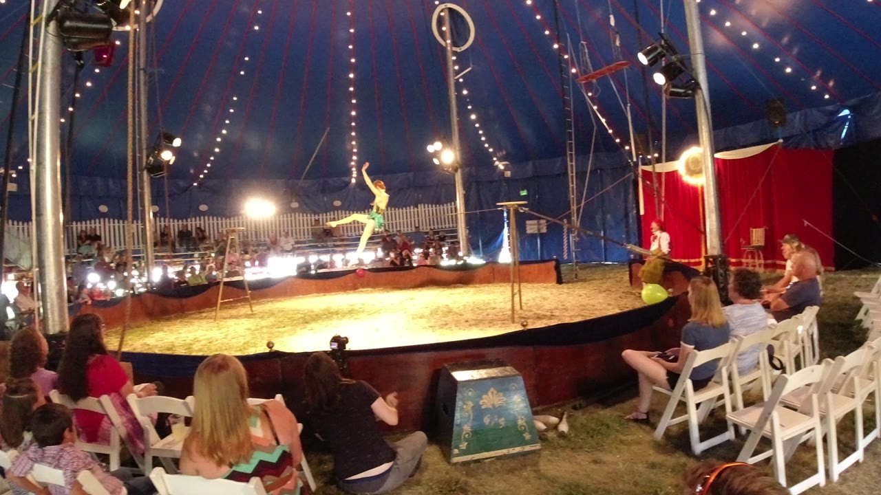 A female tightrope performer in a green outfit stands on the rope inside a Broomfield, Colorado circus tent as audience members watch from white chairs.