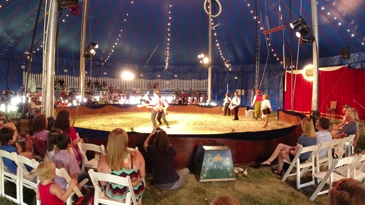 Performers continue juggling clubs in the center of a Broomfield, Colorado circus ring, with bright lights aimed across seated spectators and tent rigging above.