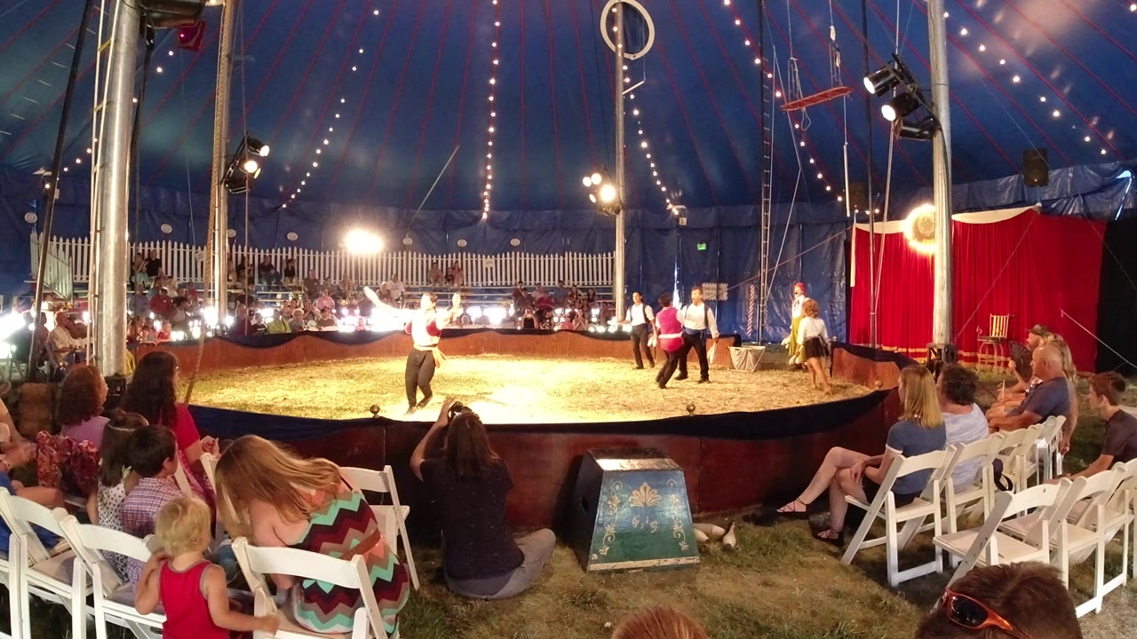 A performer in a white shirt gestures near the center of the circus ring while seated spectators and children watch from folding chairs in Broomfield, Colorado.