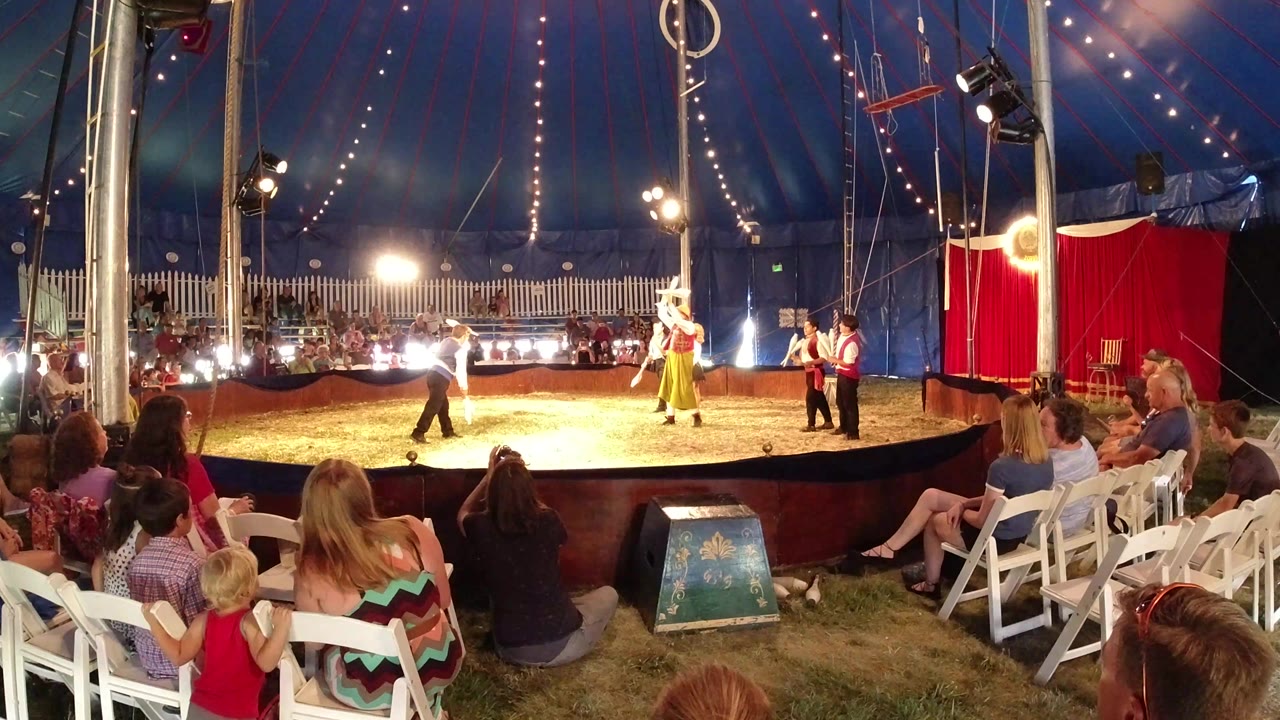Wide view of clowns performing in a circus ring as families watch from folding chairs.