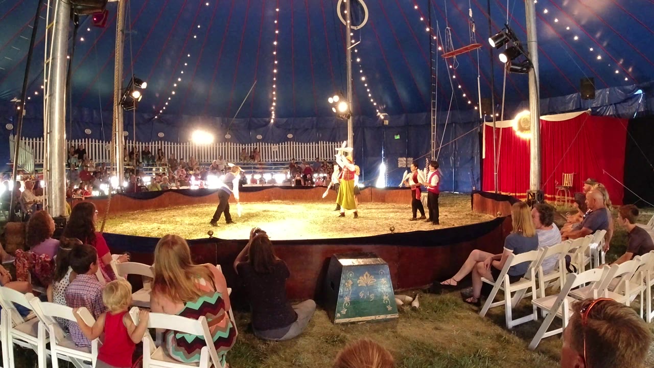 At 0:01, the Broomfield circus scene holds a wide view with the host near center, costumed clowns to the right, bright lamps behind them, and spectators in the foreground.