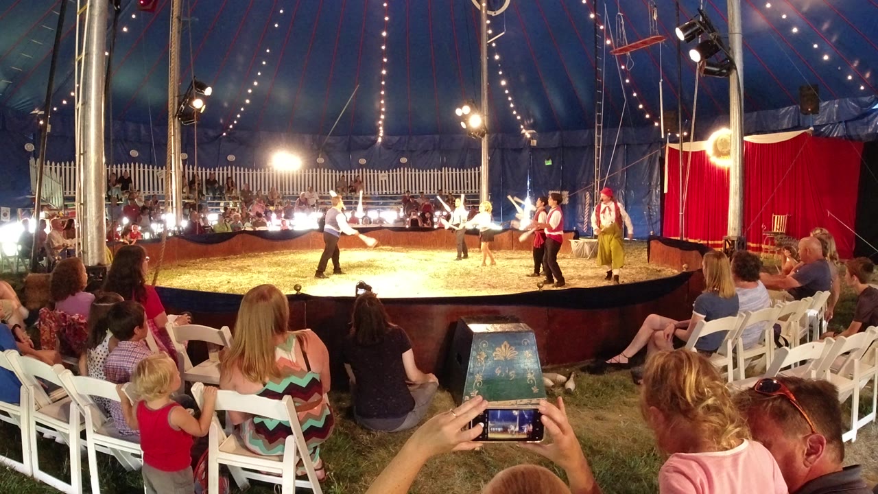Audience members and children watch costumed performers in a sawdust circus ring inside a blue tent in Broomfield, Colorado.