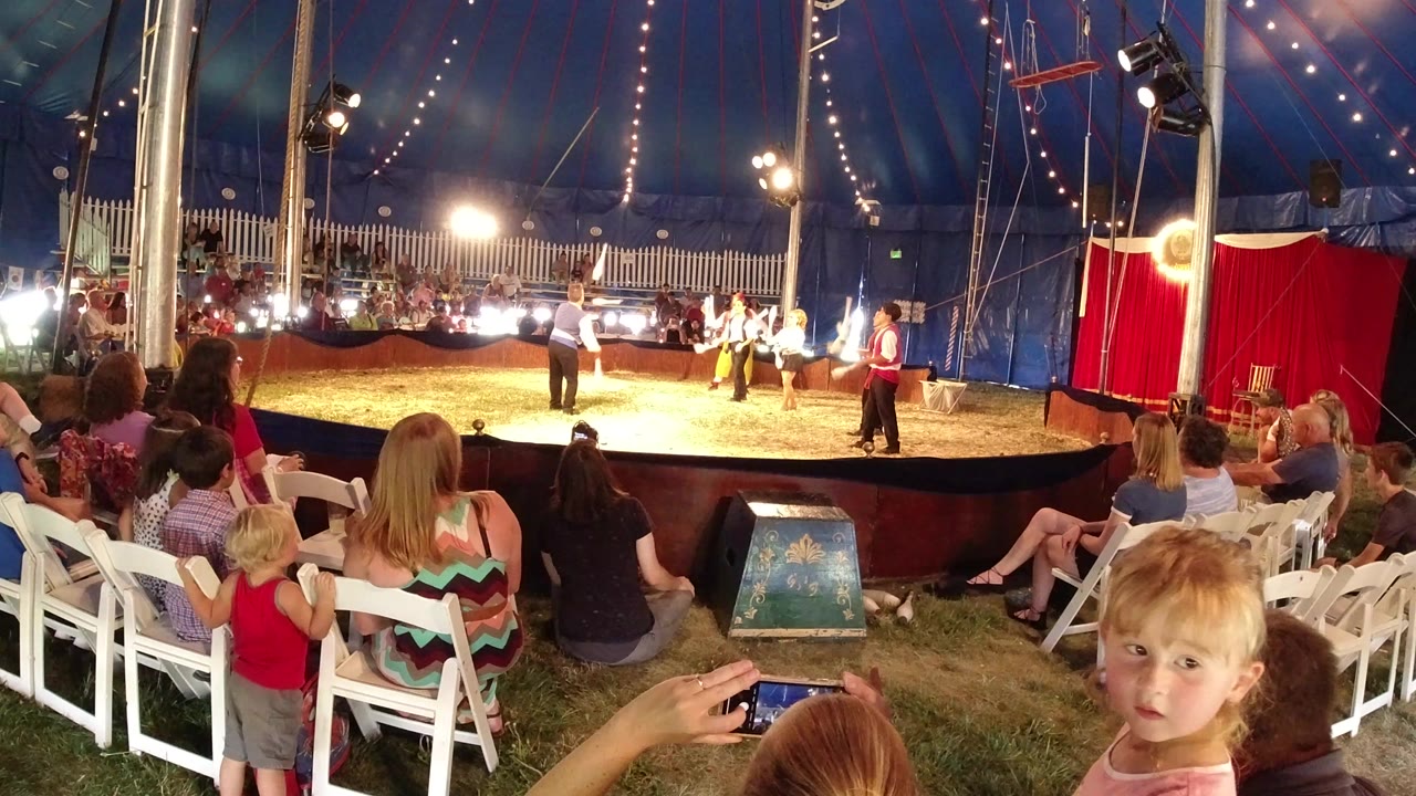 Audience members and children sit and stand near the ring in a Broomfield, Colorado circus tent as performers prepare under bright lights.