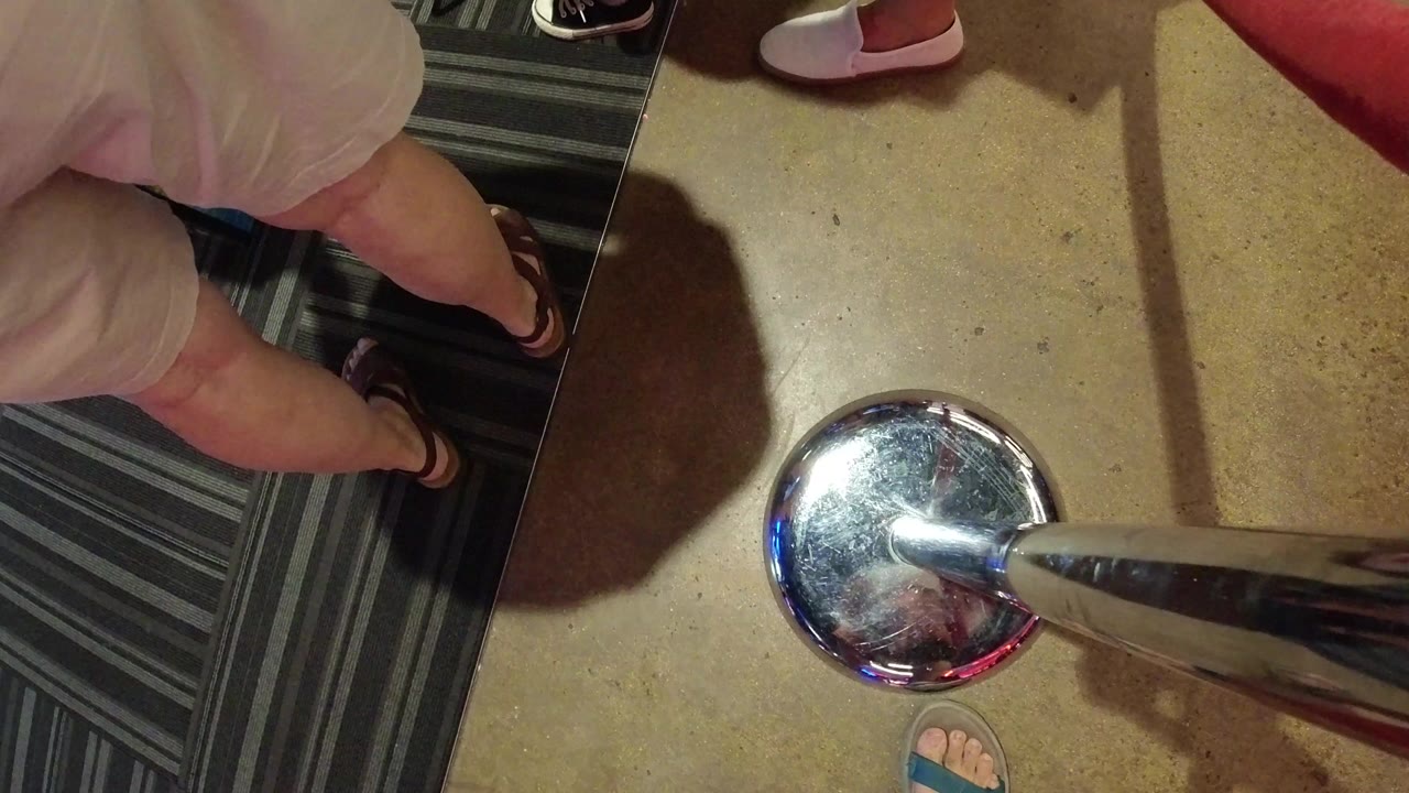 Downward view of legs, sandals, carpet, floor, and a chrome stanchion base.