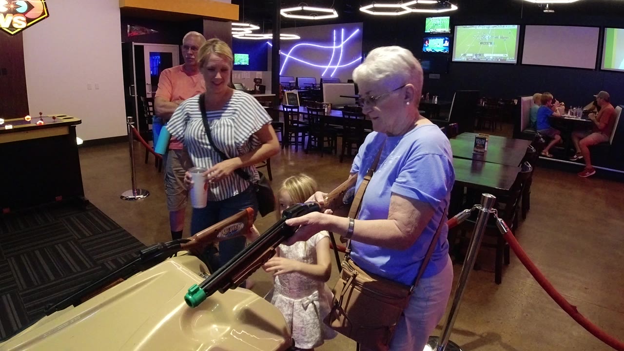 In Broomfield, Colorado, a grandmother helps or watches a young girl at a tan arcade shooting game while a blonde woman with a cup and an older man stand nearby.