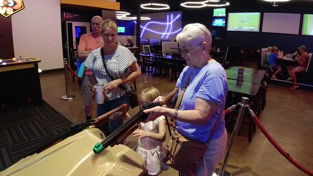 In Broomfield, Colorado, the young girl reaches over the tan arcade cabinet while her grandmother handles one of the mounted game rifles and two adults watch behind them.