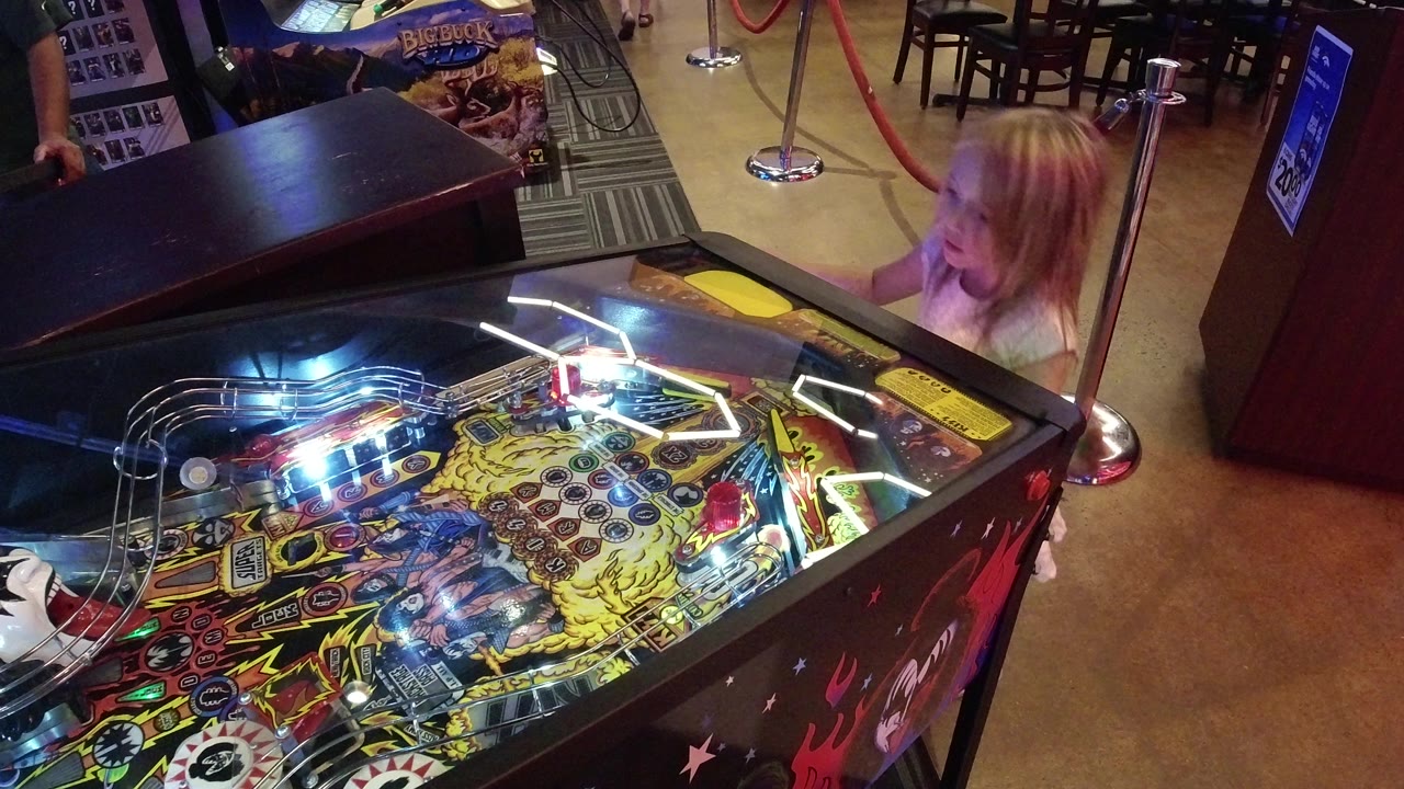 Young girl beside a lit pinball machine in an arcade room.