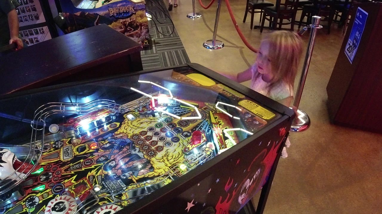 The girl remains beside the illuminated pinball table in Broomfield, Colorado, looking toward the playfield as colored lights reflect across the glass.