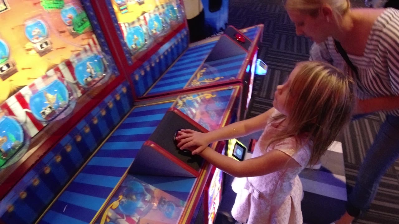 Young girl at an arcade game with an adult leaning nearby.