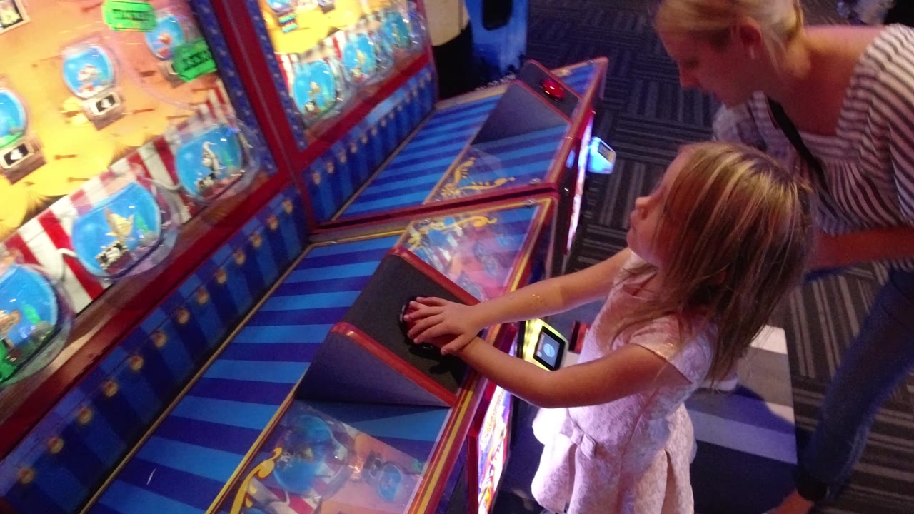 The girl reaches across the arcade console in Broomfield, Colorado as the colorful machine display glows above her and the adult stands just behind.