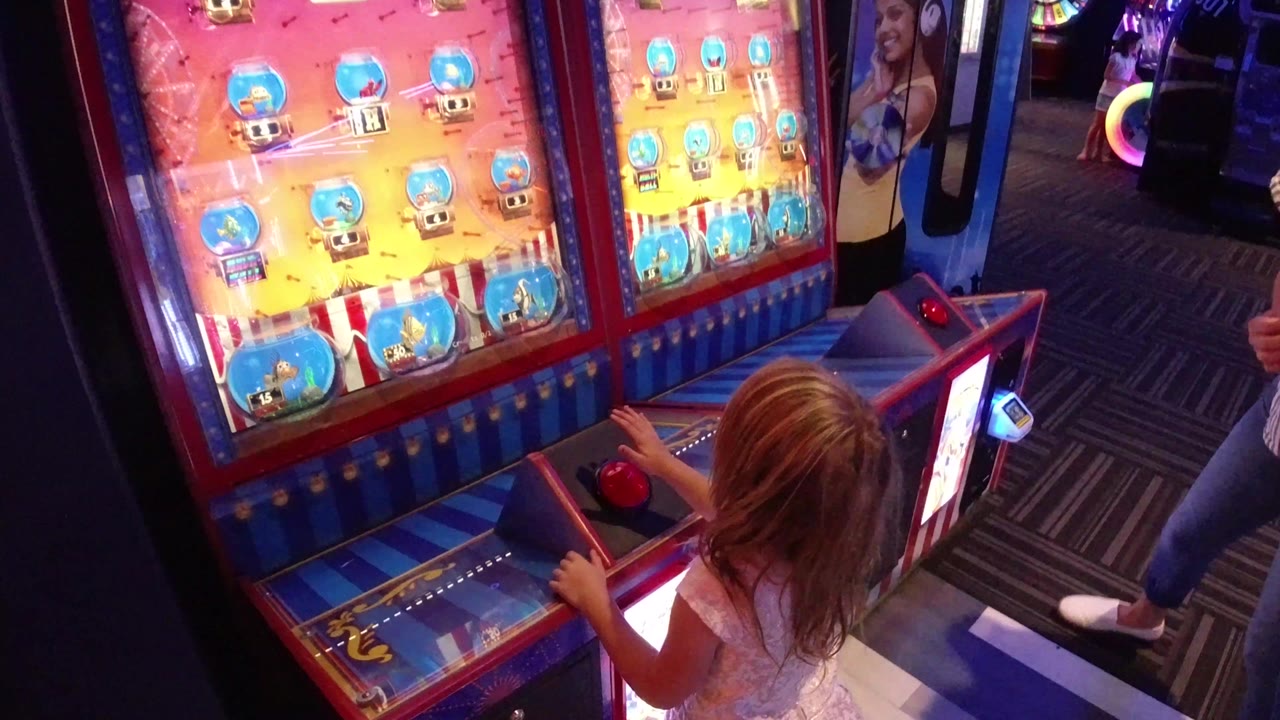 Young girl in Broomfield, Colorado stands at a bright arcade game cabinet, with the game board, red button control, patterned carpet, and nearby adult legs visible.