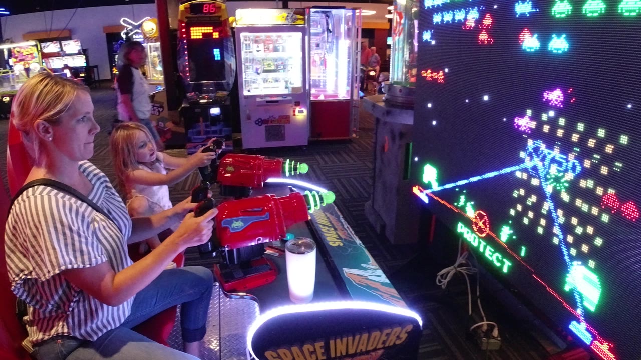 Mother and daughter play a red arcade shooter beside a glowing space game screen.