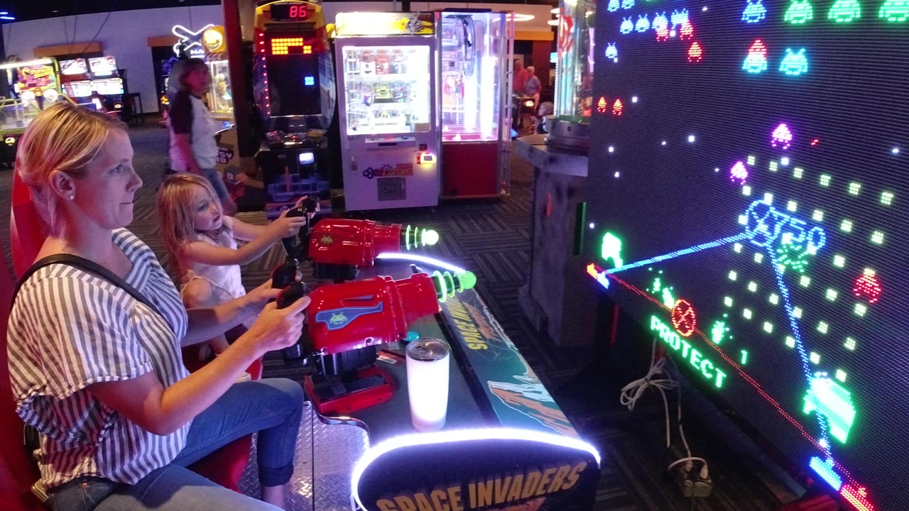 The Broomfield arcade frame shows the mother and daughter from the side as the game screen displays colored alien figures, laser lines, and the word protect.