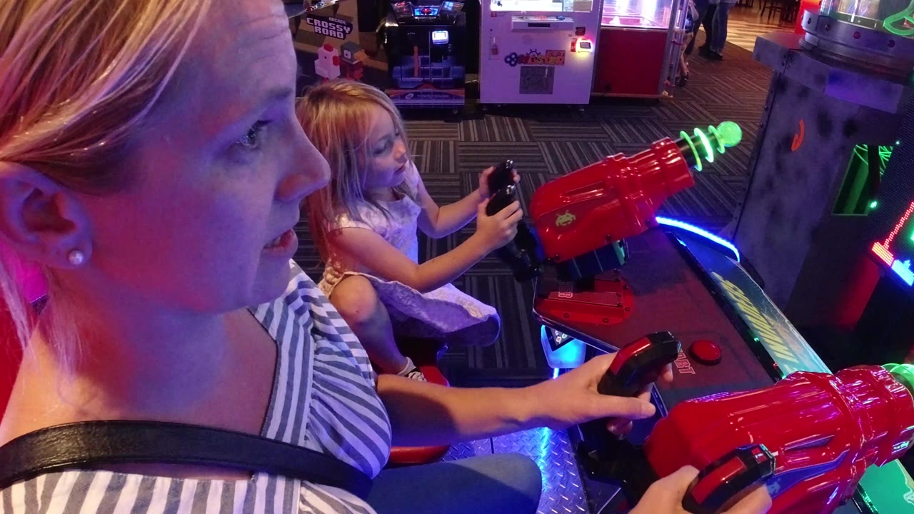 In Broomfield, Colorado, a mother in the foreground and her daughter behind her hold controls at a red arcade laser shooter under blue and pink game lights.