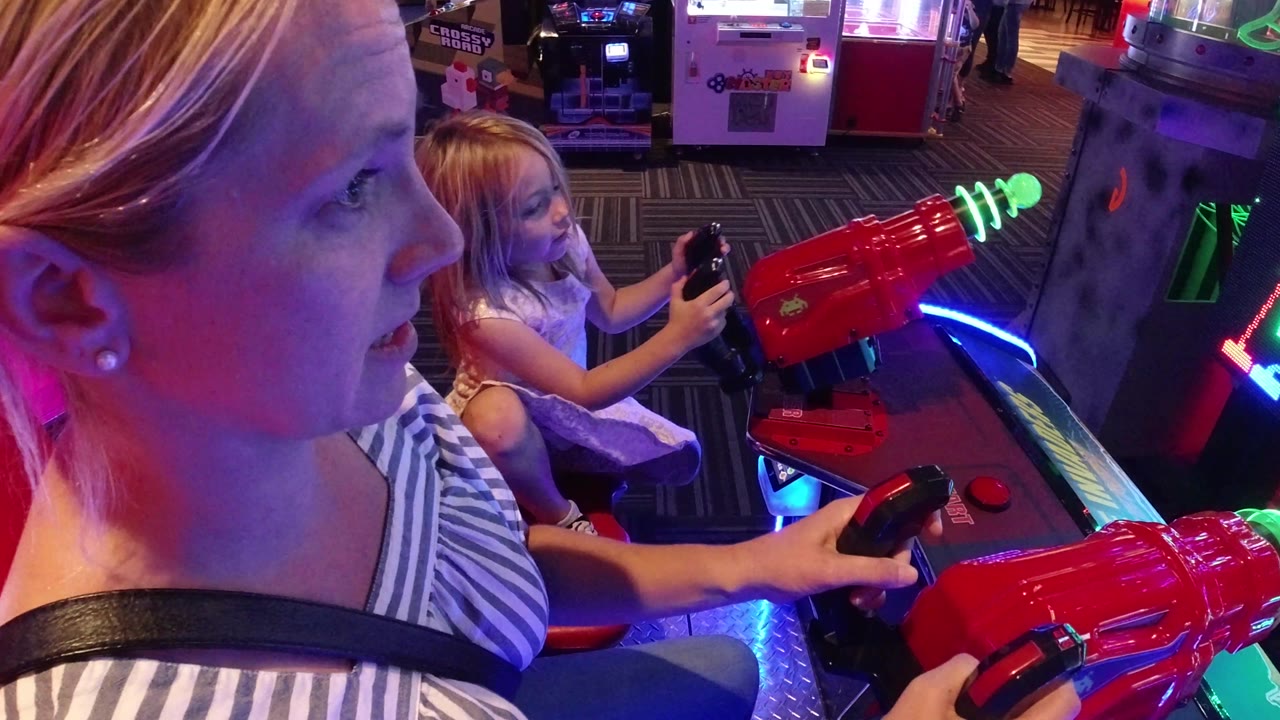 The mother looks toward the game while the daughter grips a control behind a red cannon, with glowing arcade machines visible in the background.