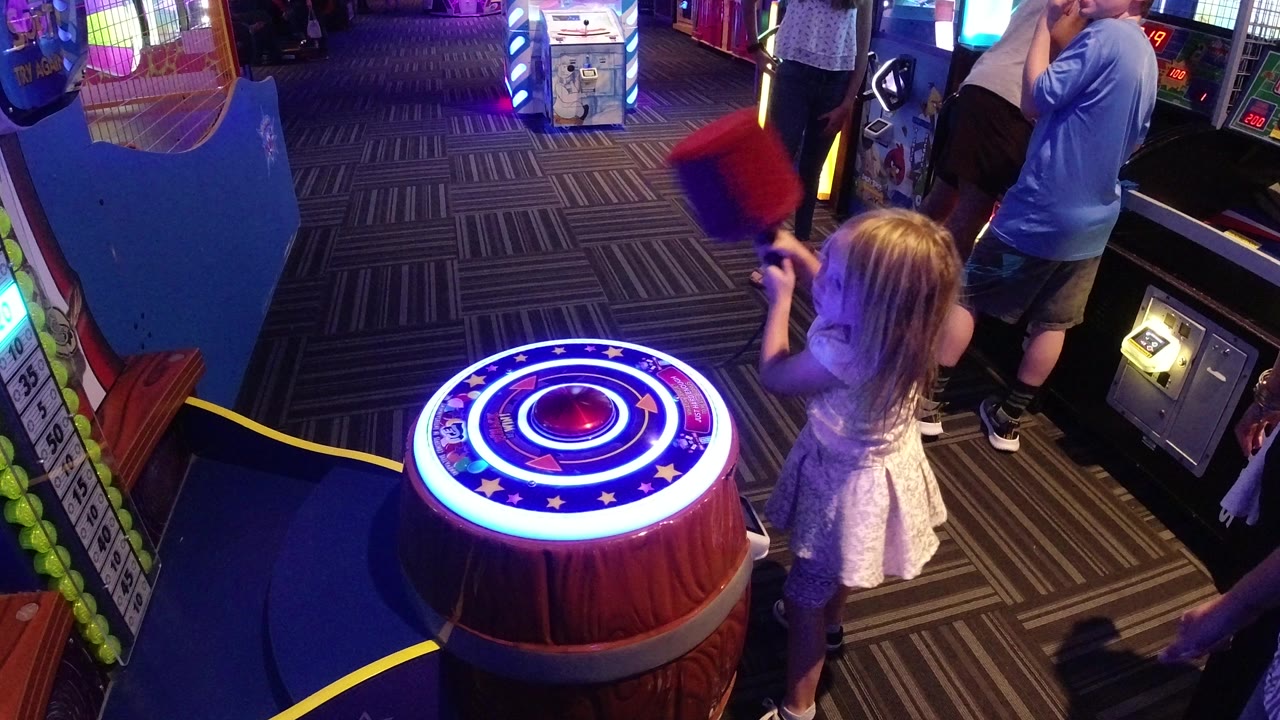 At an arcade in Broomfield, Colorado, a young girl stands beside a circular lighted hammer game while an adult woman and nearby patrons are partly visible around the machines.