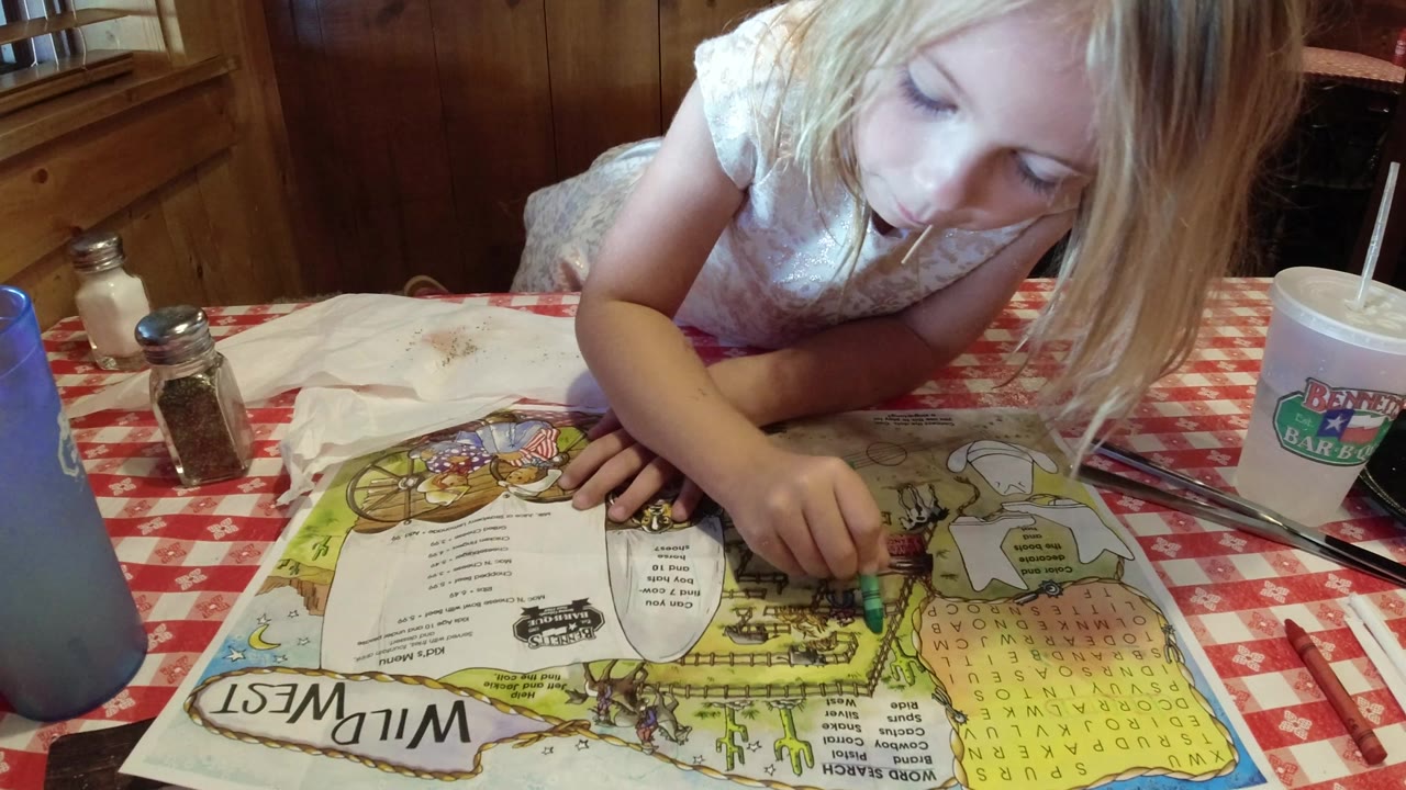 At 0:00 in Broomfield, Colorado, a young blonde girl bends over a colorful paper placemat with a green crayon beside cups and shakers on a red checkered table.
