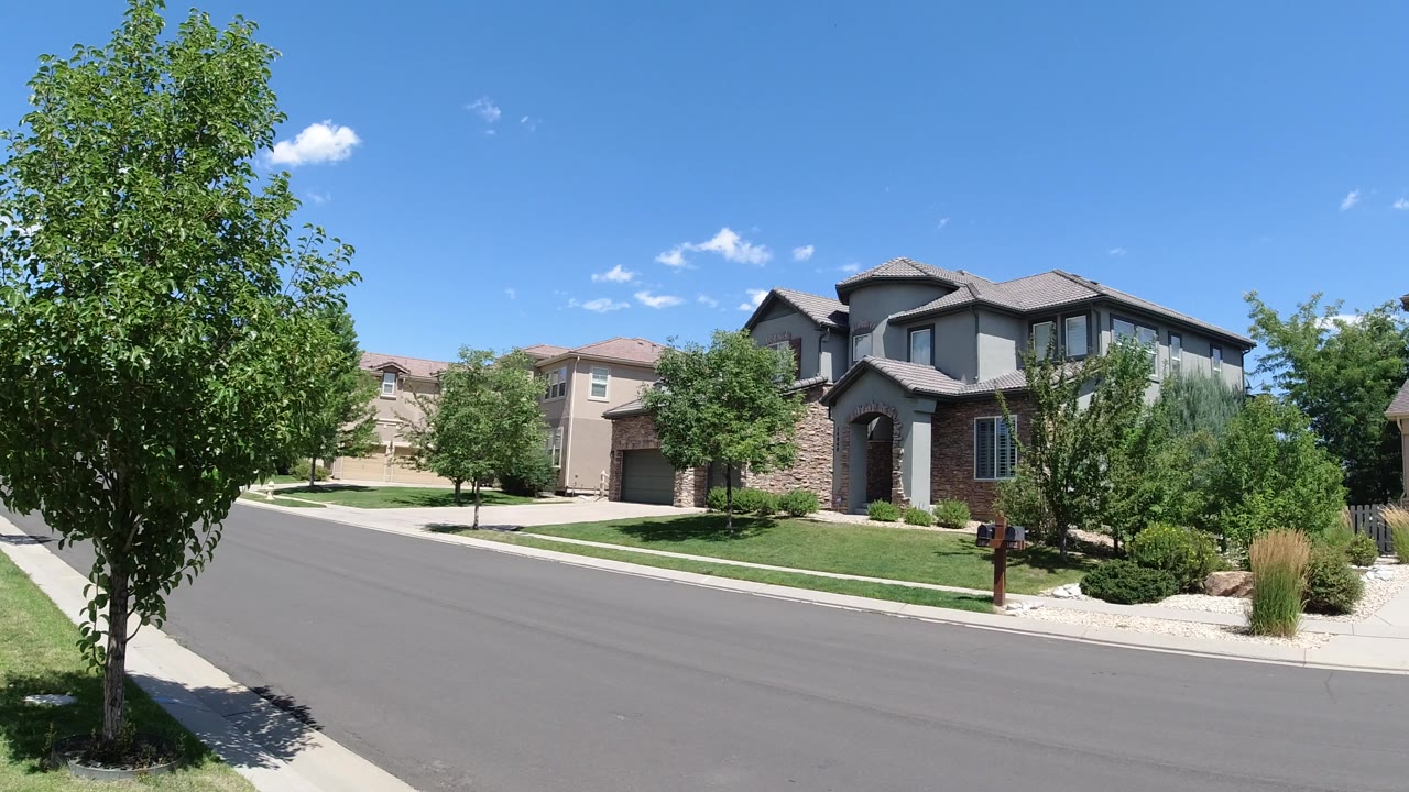 Wide view of a Broomfield, Colorado residential street with a leafy foreground tree, large two-story houses, lawns, sidewalk, and no clearly visible people.