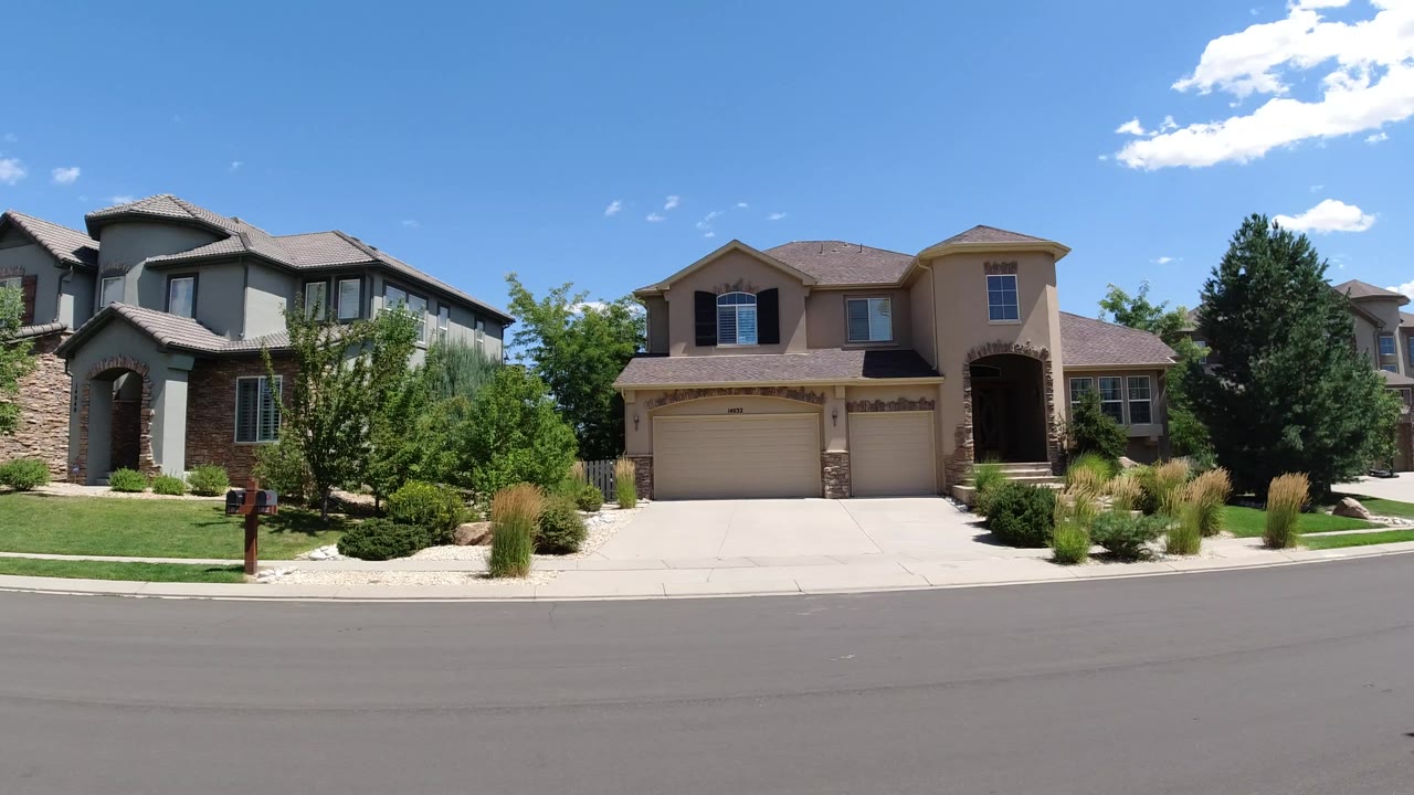 Two Broomfield homes sit behind a broad road, with trimmed greenery, driveway pavement, and bright midday sky, with no visible people.
