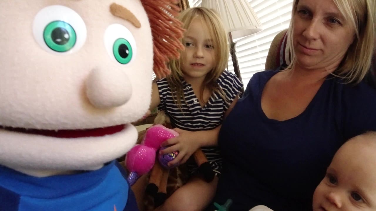 In Broomfield, Colorado, a large plush puppet fills the left foreground while a blonde child, an adult woman, and part of a baby sit in a living room.