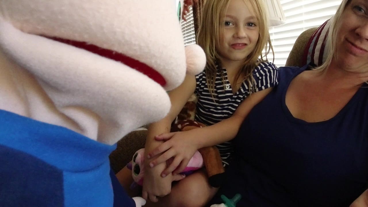 In a Broomfield, Colorado home interior, a cloth puppet fills the left foreground while a blonde woman and young girl sit together on a sofa near window blinds.