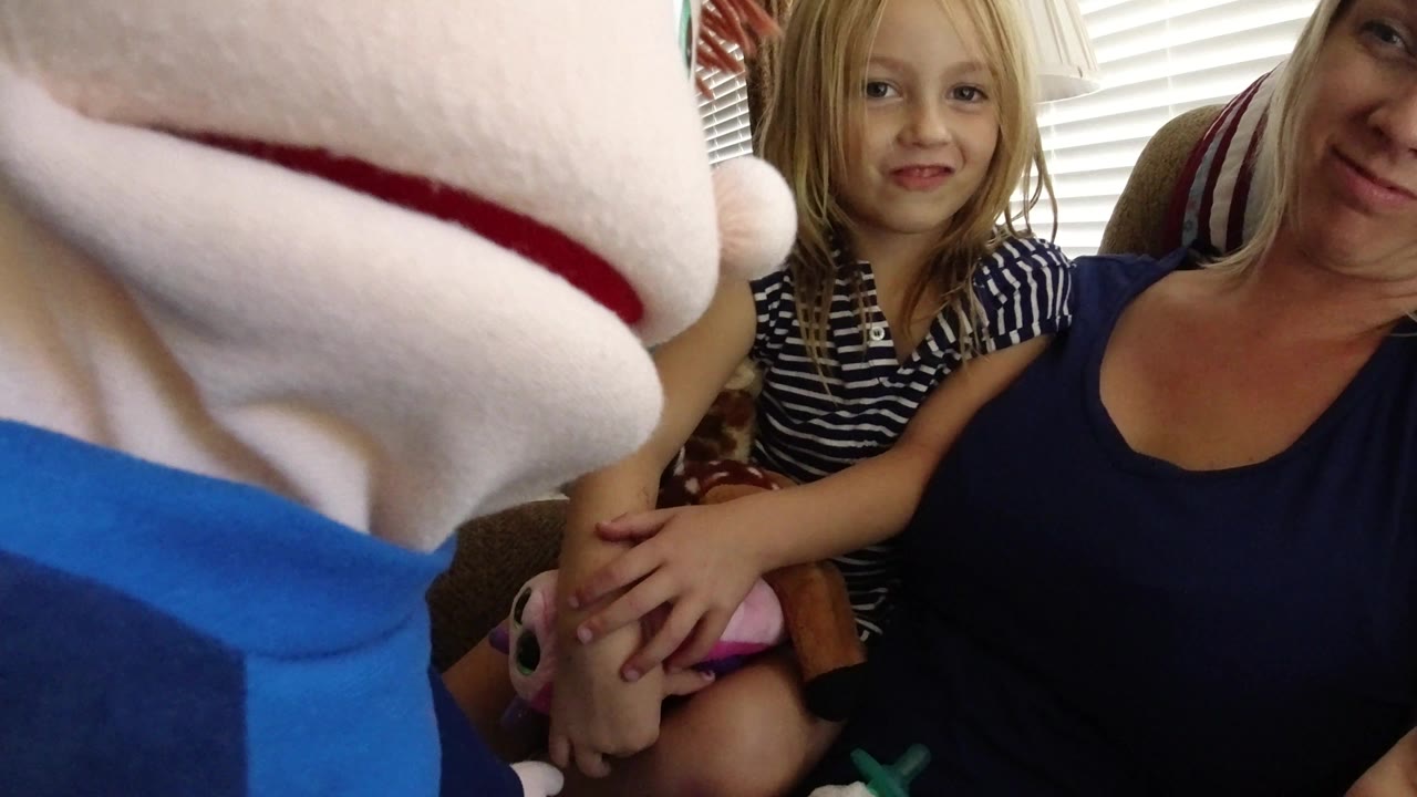 A young girl in a striped shirt sits beside a blonde woman on a sofa as the soft puppet remains close to the lens inside the Broomfield, Colorado home.