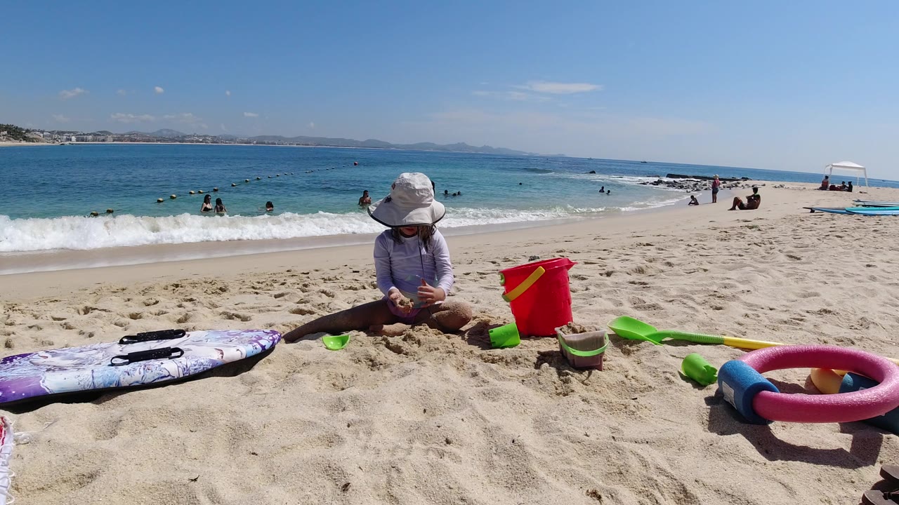 Child sitting in beach sand beside toys and ocean waves.